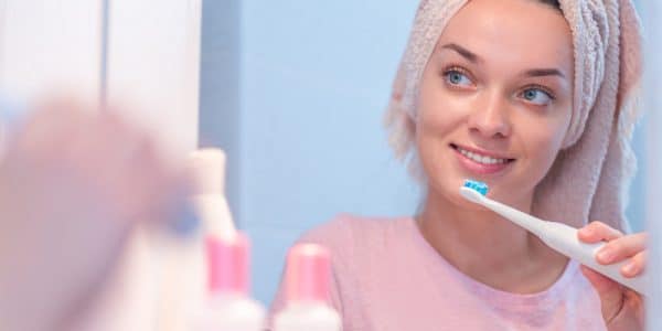 Woman looking in the mirror and brushing her teeth with an electric toothbrush to maintain oral health.