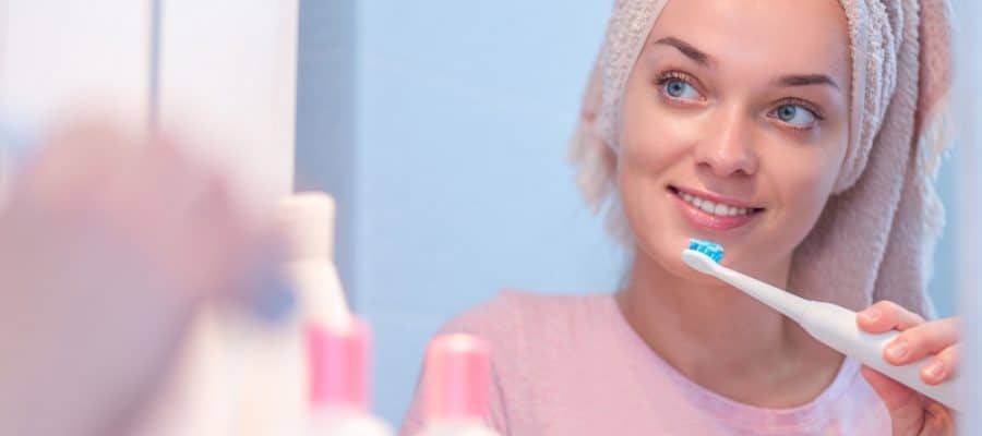 Woman looking in the mirror and brushing her teeth with an electric toothbrush to maintain oral health.